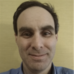 A person with short, dark hair and a slight smile is facing the camera. They are wearing a blue shirt and have a neutral background