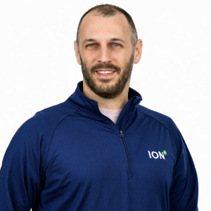 A man with a beard and short hair smiles while wearing a navy blue zip-up jacket with the logo "ION" on the left side. The background is plain and white