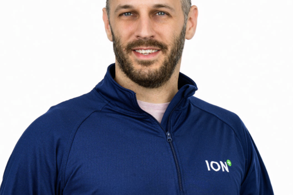 A man with a beard and short hair smiles while wearing a navy blue zip-up jacket with the logo "ION" on the left side. The background is plain and white