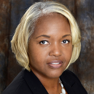 A woman with short, wavy blonde hair and a light complexion is looking directly at the camera. She is wearing a black blazer and a silver necklace. The background is a blurred, neutral-toned fabric