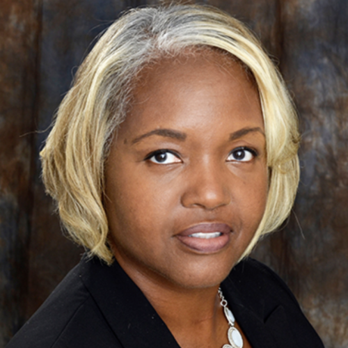 A woman with short, wavy blonde hair and a light complexion is looking directly at the camera. She is wearing a black blazer and a silver necklace. The background is a blurred, neutral-toned fabric