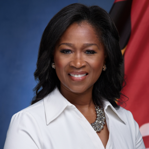 A woman with long, dark hair smiles at the camera, wearing a white blouse and a statement necklace. The background features a blue gradient and a flag