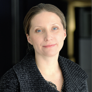 A woman with light brown hair and blue eyes is smiling softly. She is wearing a dark, textured sweater and is positioned in a well-lit indoor setting with a blurred background