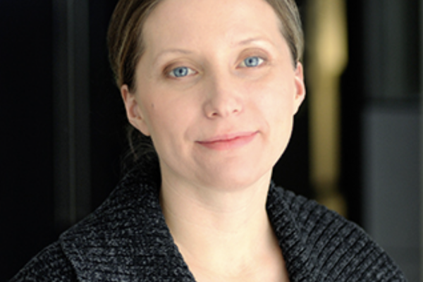 A woman with light brown hair and blue eyes is smiling softly. She is wearing a dark, textured sweater and is positioned in a well-lit indoor setting with a blurred background