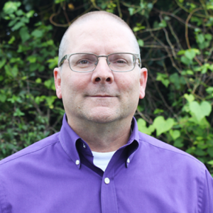 A man with short hair and glasses stands outdoors, wearing a purple button-up shirt. He has a neutral expression and is positioned in front of a background of green foliage