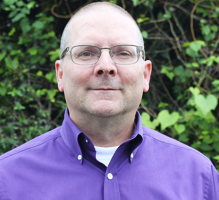 A man with short hair and glasses stands outdoors, wearing a purple button-up shirt. He has a neutral expression and is positioned in front of a background of green foliage