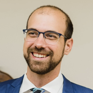 A man with a short beard and glasses smiles while wearing a blue suit and a white shirt with a patterned tie. He has a bald head with some hair on the sides. The background is neutral and blurred