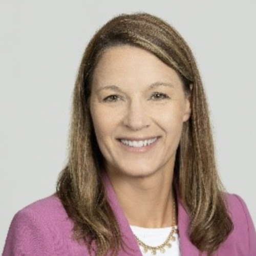 A woman with long, straight brown hair smiles at the camera. She is wearing a pink blazer and a white top, with a delicate necklace. The background is light gray