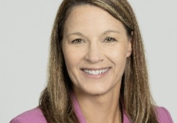 A woman with long, straight brown hair smiles at the camera. She is wearing a pink blazer and a white top, with a delicate necklace. The background is light gray