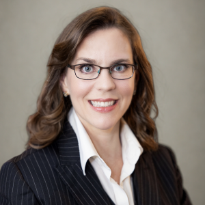 A woman with long, wavy brown hair and glasses smiles at the camera. She is wearing a dark pinstripe suit jacket over a white blouse. The background is a soft, neutral color
