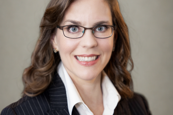 A woman with long, wavy brown hair and glasses smiles at the camera. She is wearing a dark pinstripe suit jacket over a white blouse. The background is a soft, neutral color