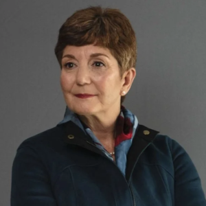 A woman with short brown hair and a subtle smile is wearing a dark blue jacket and a patterned scarf. She is facing slightly to the side against a gray background
