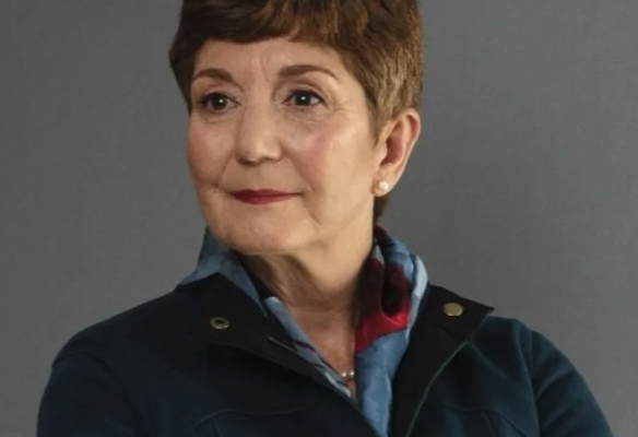 A woman with short brown hair and a subtle smile is wearing a dark blue jacket and a patterned scarf. She is facing slightly to the side against a gray background