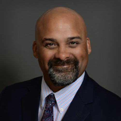 A man with a bald head and a neatly trimmed beard smiles at the camera. He is wearing a dark suit with a light blue dress shirt and a patterned tie. The background is a neutral gray