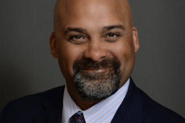A man with a bald head and a neatly trimmed beard smiles at the camera. He is wearing a dark suit with a light blue dress shirt and a patterned tie. The background is a neutral gray
