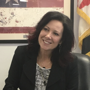 A woman with shoulder-length dark hair and a slight smile is seated at a desk. She is wearing a black blazer over a patterned top and has earrings. In the background, there is a framed image with a star and a flag