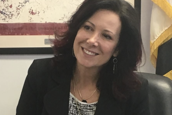 A woman with shoulder-length dark hair and a slight smile is seated at a desk. She is wearing a black blazer over a patterned top and has earrings. In the background, there is a framed image with a star and a flag