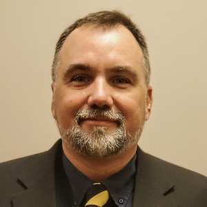 A man with a beard and short hair is wearing a black suit jacket over a dark shirt and a patterned tie. He is smiling and looking directly at the camera. The background is a plain light color