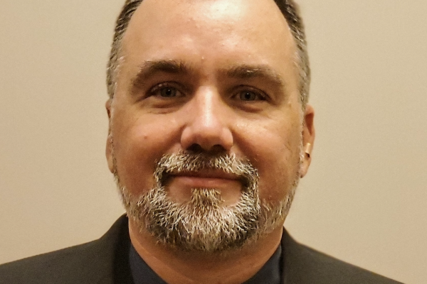 A man with a beard and short hair is wearing a black suit jacket over a dark shirt and a patterned tie. He is smiling and looking directly at the camera. The background is a plain light color