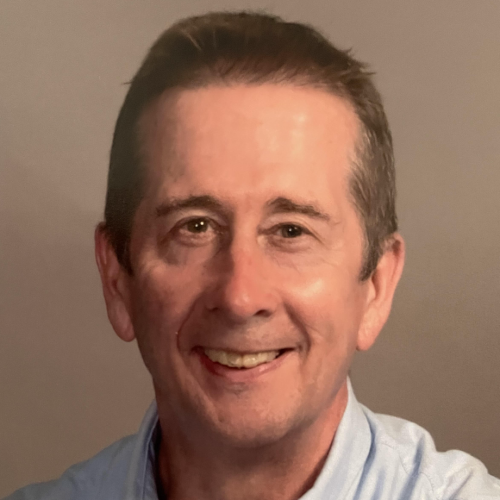 A middle-aged man with short, light brown hair and a friendly smile is looking directly at the camera. He is wearing a light blue collared shirt. The background is a neutral color