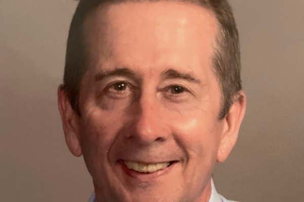 A middle-aged man with short, light brown hair and a friendly smile is looking directly at the camera. He is wearing a light blue collared shirt. The background is a neutral color