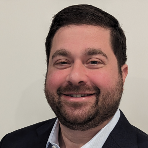 A man with a short beard and dark hair smiles at the camera. He is wearing a dark blazer over a light-colored shirt. The background is plain and light-colored