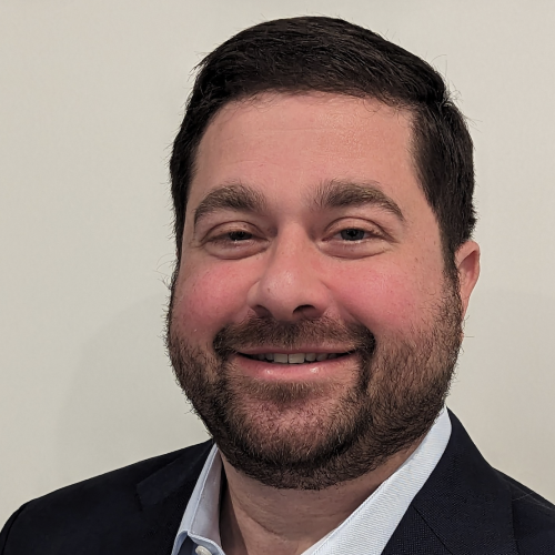 A man with a short beard and dark hair smiles at the camera. He is wearing a dark blazer over a light-colored shirt. The background is plain and light-colored
