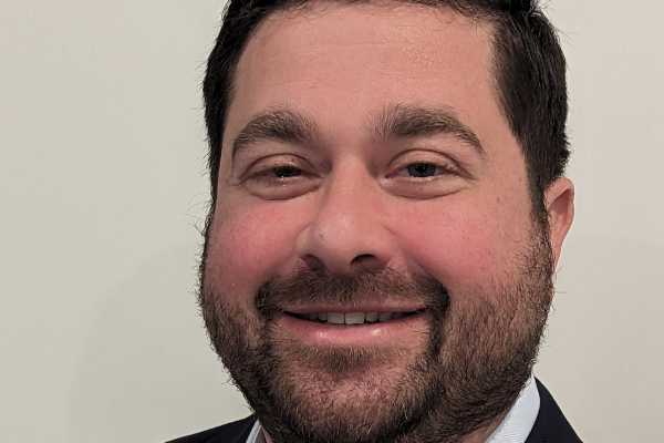 A man with a short beard and dark hair smiles at the camera. He is wearing a dark blazer over a light-colored shirt. The background is plain and light-colored