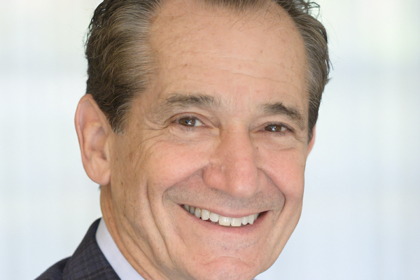 A middle-aged man with short, dark hair and a friendly smile is wearing a dark blazer over a light shirt. He is facing the camera against a softly blurred background