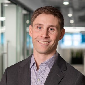 A man with short brown hair and a friendly smile is wearing a dark suit jacket over a light checkered shirt. He is standing in an office environment with glass walls and a bright background