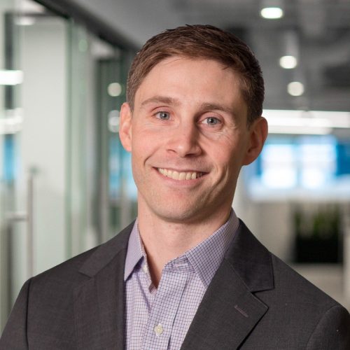 A man with short brown hair and a friendly smile is wearing a dark suit jacket over a light checkered shirt. He is standing in an office environment with glass walls and a bright background
