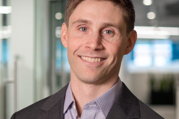 A man with short brown hair and a friendly smile is wearing a dark suit jacket over a light checkered shirt. He is standing in an office environment with glass walls and a bright background