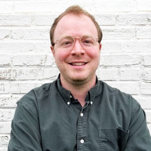 A smiling person with short, light brown hair and glasses is sitting against a white brick wall. They are wearing a dark button-up shirt and have a relaxed expression
