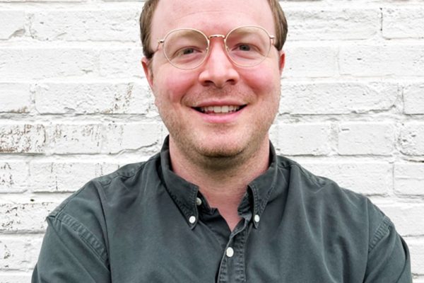 A smiling person with short, light brown hair and glasses is sitting against a white brick wall. They are wearing a dark button-up shirt and have a relaxed expression