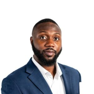 A man with a short beard and closely cropped hair is wearing a dark blue suit jacket over a white shirt. He is looking directly at the camera with a neutral expression against a plain white background