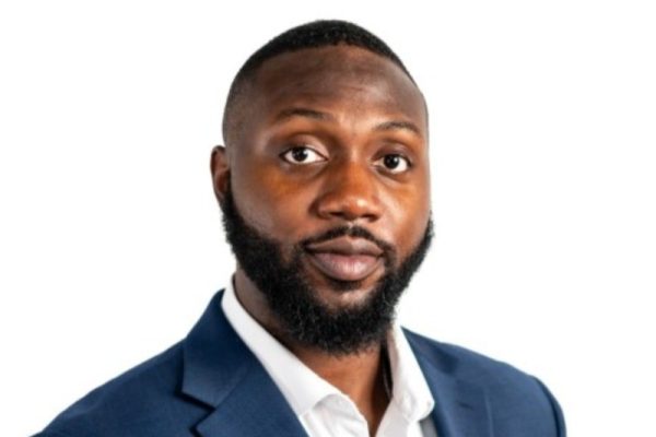 A man with a short beard and closely cropped hair is wearing a dark blue suit jacket over a white shirt. He is looking directly at the camera with a neutral expression against a plain white background