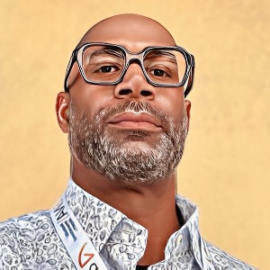 A man with a short beard and glasses poses confidently, wearing a patterned shirt and a lanyard around his neck. The background is a solid light color