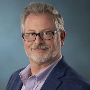 A middle-aged man with gray hair and a beard smiles while wearing glasses. He is dressed in a light purple shirt and a dark blue blazer, set against a soft blue background