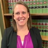 A woman with shoulder-length brown hair smiles while wearing a black blazer over a purple blouse. She stands in front of a bookshelf filled with green and red legal books