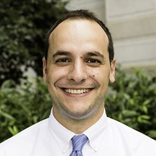 A man with short brown hair and a friendly smile is wearing a white dress shirt and a light blue tie. He is standing outdoors in front of greenery and a stone wall