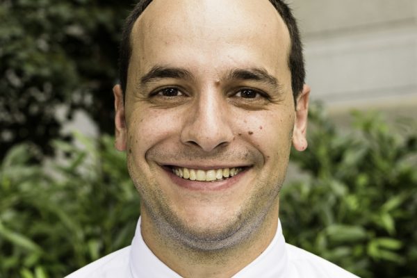A man with short brown hair and a friendly smile is wearing a white dress shirt and a light blue tie. He is standing outdoors in front of greenery and a stone wall