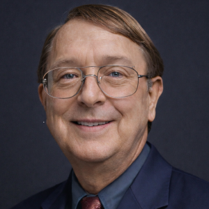 A middle-aged man with short brown hair and glasses smiles at the camera. He is wearing a dark suit with a light blue shirt and a maroon tie. The background is dark