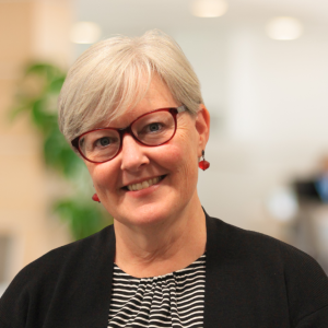 A woman with short gray hair and glasses smiles at the camera. She is wearing a black cardigan over a striped shirt and has red earrings. The background is blurred, suggesting an office environment