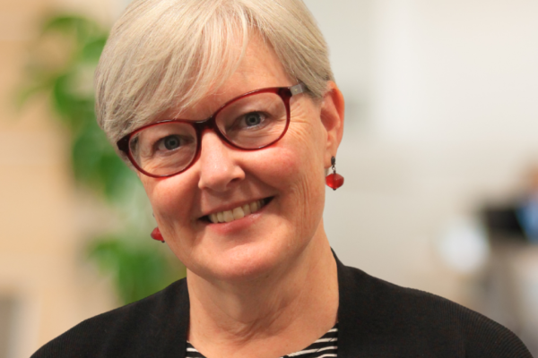 A woman with short gray hair and glasses smiles at the camera. She is wearing a black cardigan over a striped shirt and has red earrings. The background is blurred, suggesting an office environment