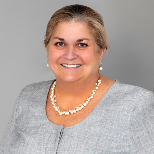 A woman with shoulder-length light brown hair smiles at the camera. She is wearing a gray blouse and a pearl necklace. The background is a plain light gray