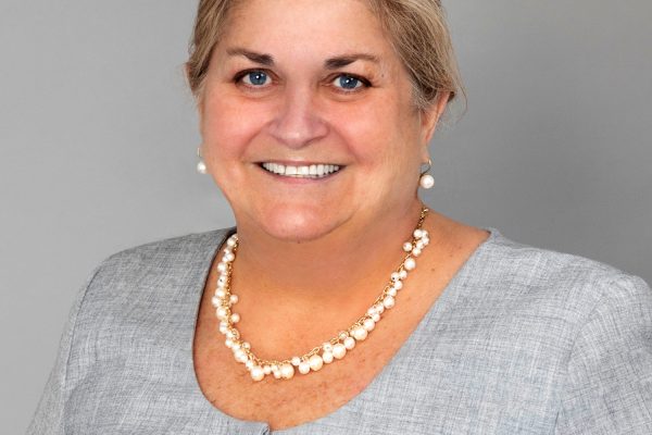 A woman with shoulder-length light brown hair smiles at the camera. She is wearing a gray blouse and a pearl necklace. The background is a plain light gray