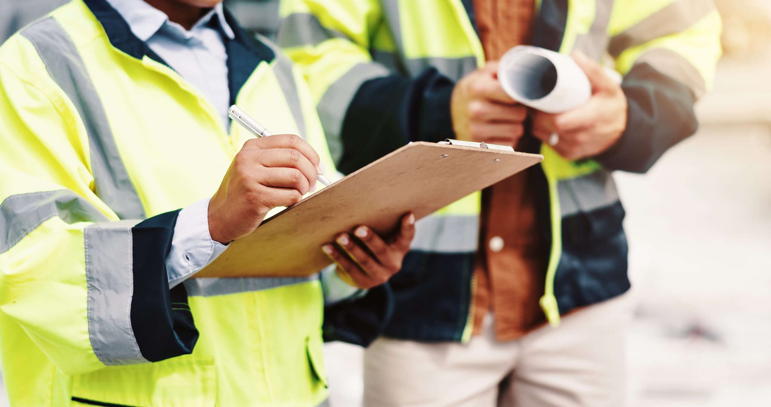 Two construction workers are on-site. One worker, wearing a bright yellow safety jacket, is holding a clipboard and writing with a pen. The other worker, dressed in a black jacket, is holding a rolled-up blueprint