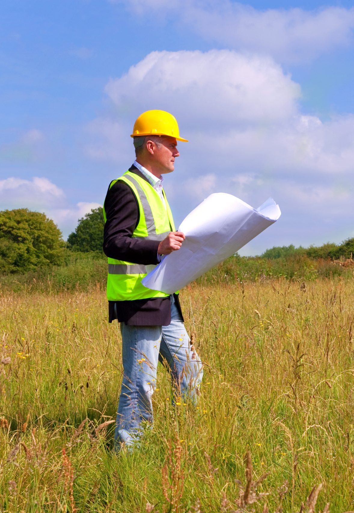 A person wearing a yellow hard hat and a reflective safety vest stands in a grassy field, holding a set of blueprints. The background features trees and a clear blue sky with some clouds