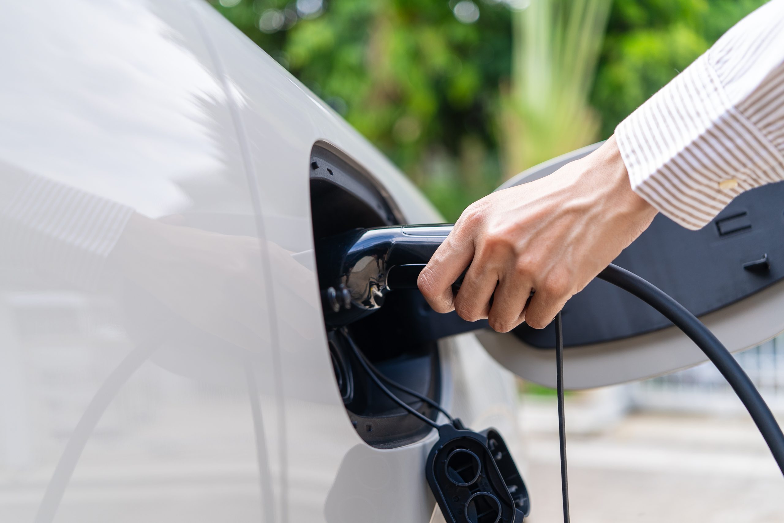 A person is plugging an electric vehicle charging cable into the charging port of a car. The background features greenery, indicating an outdoor setting