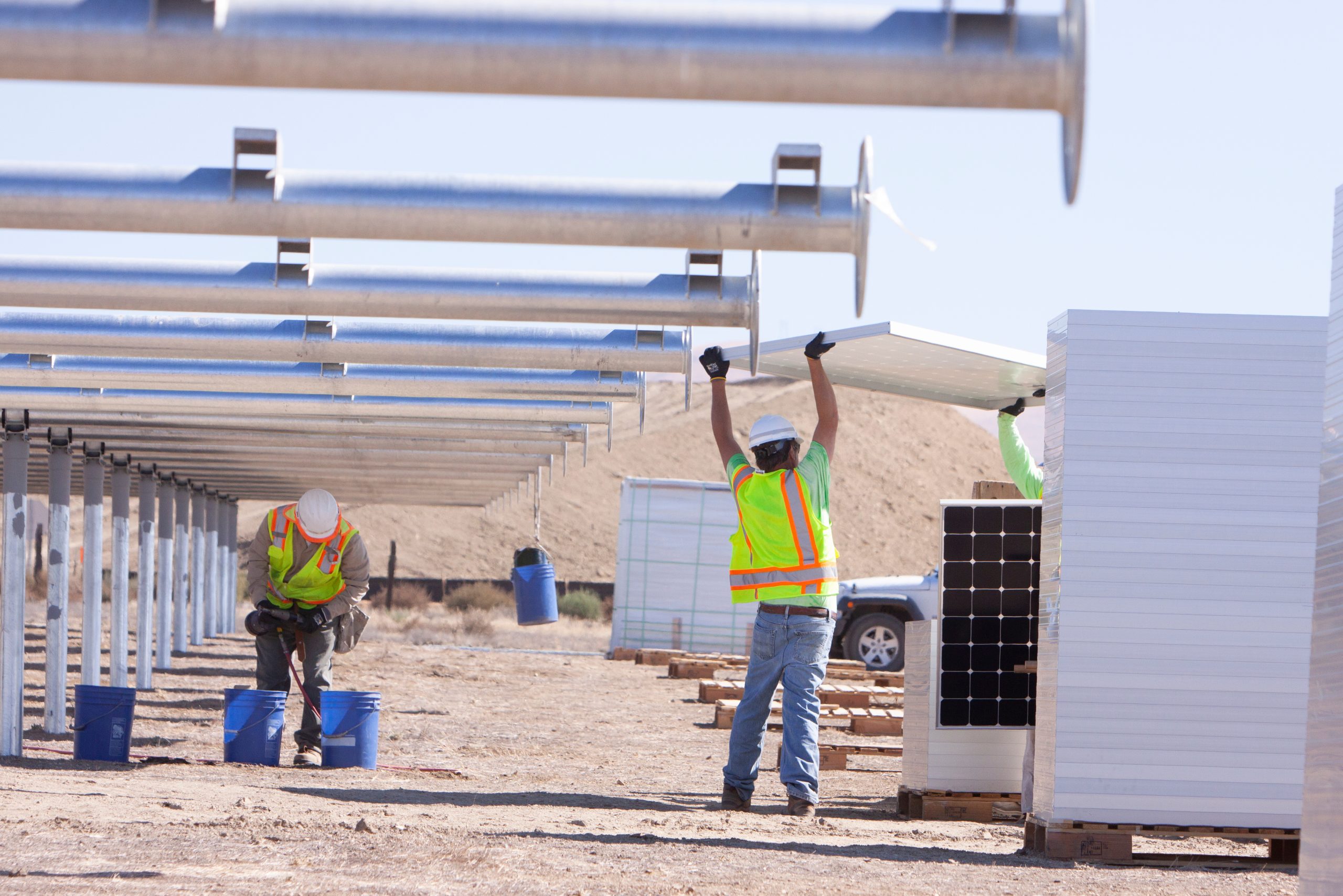 Two workers in safety vests are installing solar panels on a construction site. One worker is lifting a solar panel overhead, while the other is positioned nearby, preparing additional panels. In the background, there are stacks of solar panels and a vehicle parked on the site. The landscape is arid with a few hills in the distance
